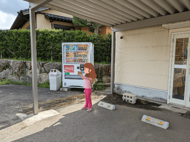 佐賀県伊万里市にある「りんちゃん牧場ふれあいパーク」近くにある自販機の前で女の子がジュースを飲んでいる画像
