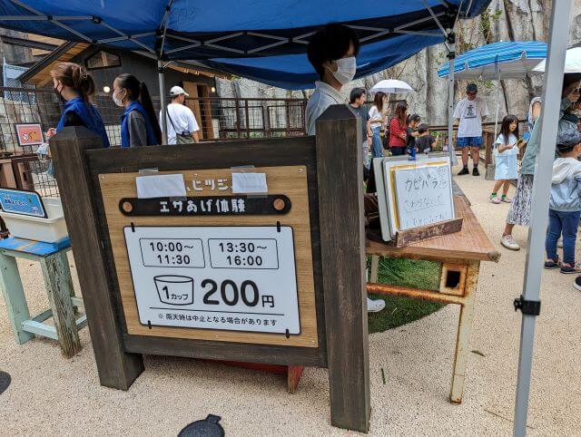 沖縄県沖縄市胡屋にある動物園「こどもの国沖縄ズージアム」のヒツジの餌あげ体験の様子。1カップ200円と営業時間が書いてある。