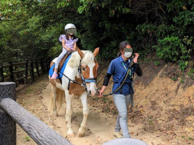 福岡県北九州市にある「響灘緑地グリーンパーク」の乗馬体験をしているところ。スタッフの女性が馬をひいていて女の子が馬の背中に乗っている画像。