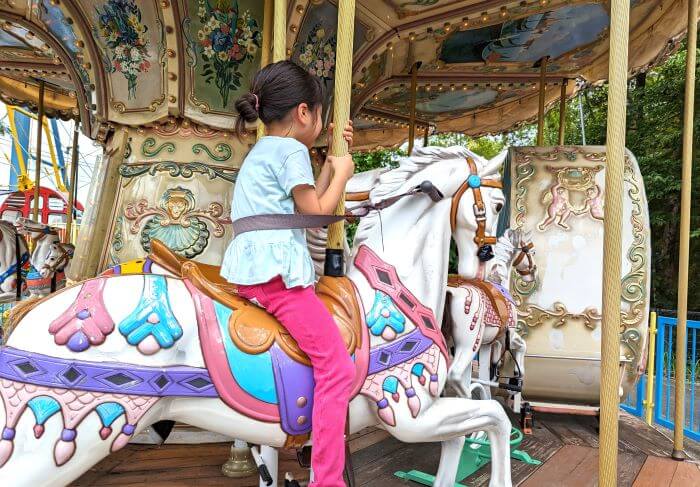 福岡市動物園の南園に位置する遊戯施設「ミニ遊園地」のメリーゴーランドの白馬に6歳の女の子がシートベルトをして乗っている画像。